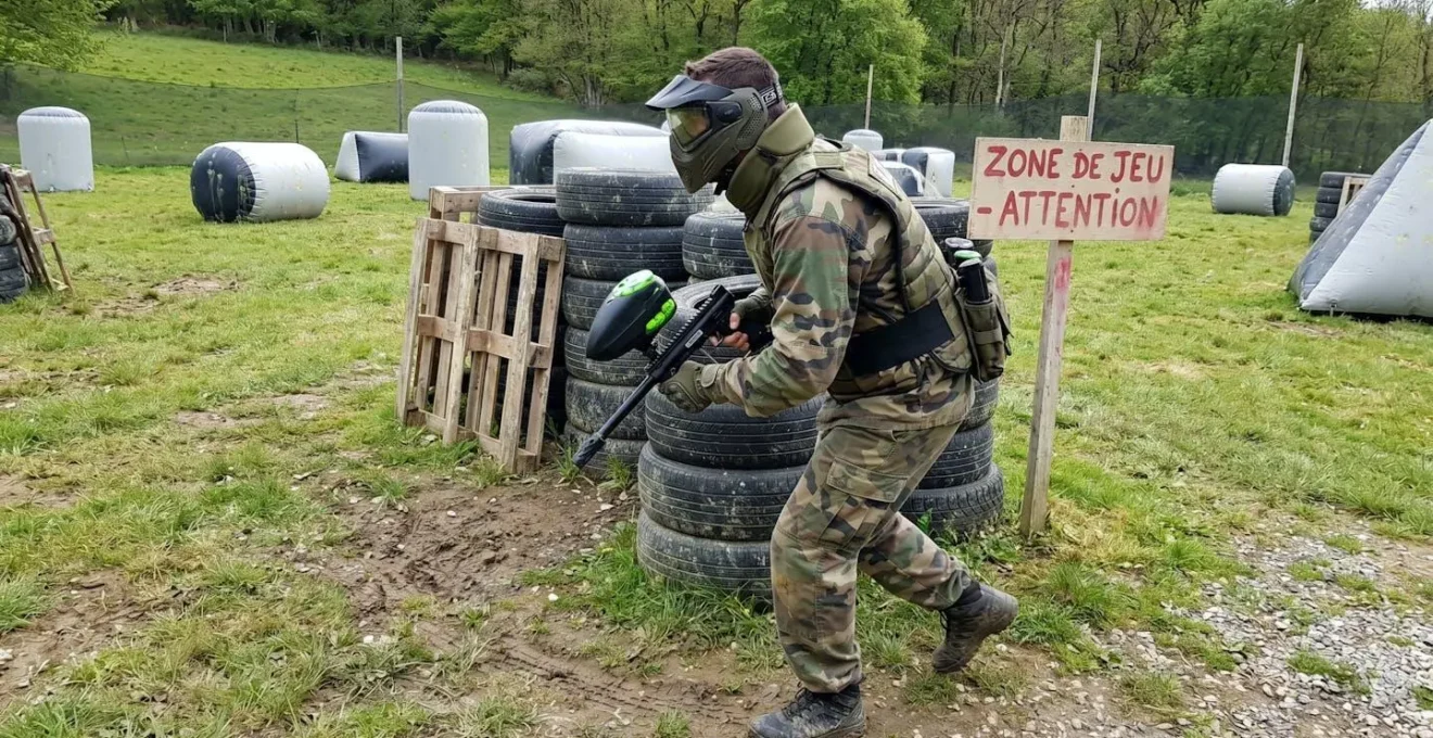 Un joueur de paintball vu de dos en position dynamique sur un terrain outdoor, portant un masque de protection intégral bien visible, avec des structures gonflables en arrière-plan