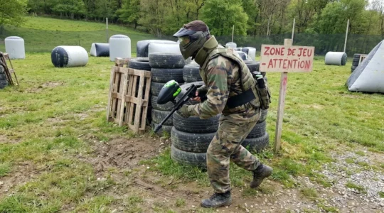 Un joueur de paintball vu de dos en position dynamique sur un terrain outdoor, portant un masque de protection intégral bien visible, avec des structures gonflables en arrière-plan